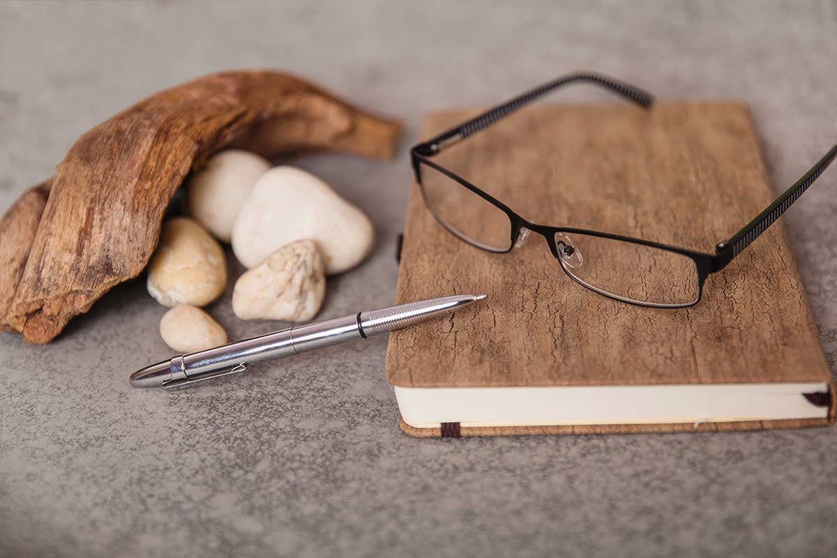 Brille auf einem Notizbuch, daneben Kugelschreiber, Kieselsteine und Treibholz
